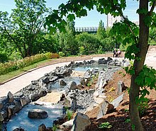 Wasserlauf, von einem kleinen Teich ausgehend, mit 2 Stufen abwärts. Der Wasserlauf ist gesäumt von groben  Natursteinen, darum ein barrierefreier Weg. Am Teich sitzen 2 Bewohner auf einer Bank, die Umgebung ist bewaldet